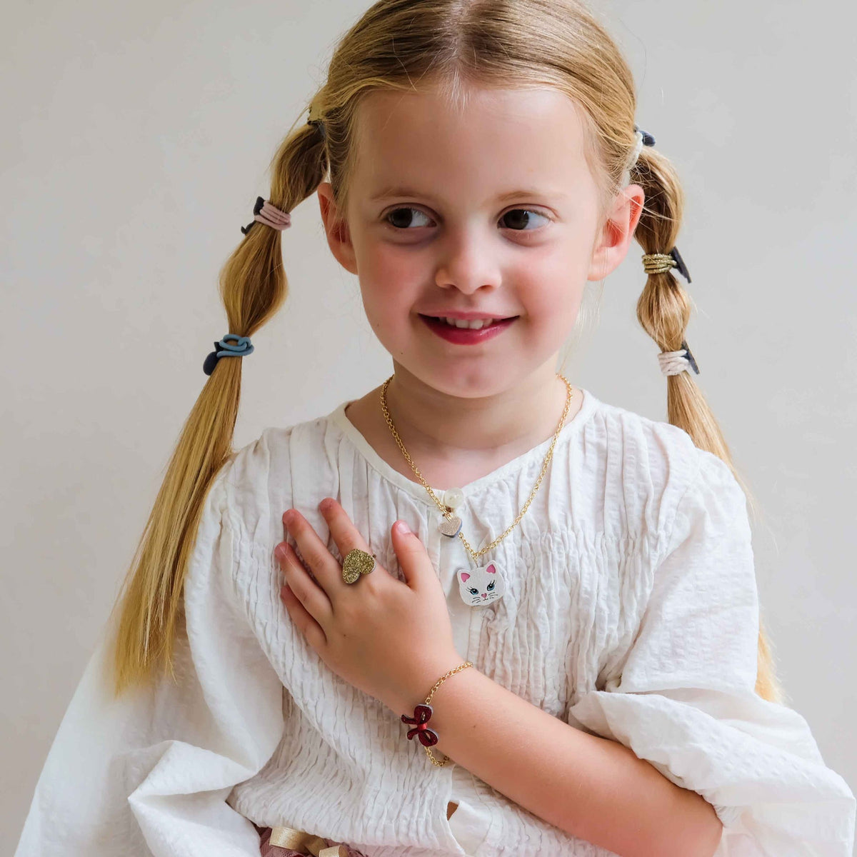 A young girl with blonde pigtails wearing a white textured blouse and gold jewelry, including a cat-shaped pendant necklace, a ring, and a bracelet, posing against a light neutral background.