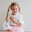 A young girl with blonde pigtails smiles while wearing a white textured blouse and a pink tulle skirt with small red bow prints, posed against a plain light gray background.