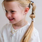 A young girl with blonde hair styled in a side ponytail wearing a gold chain necklace with a cat-shaped pendant and matching hair accessories, against a plain light gray background.