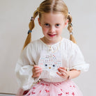A young girl with blonde pigtails holds a white cat-shaped sticker sheet decorated with colorful hearts, stars, and a gold heart. She is wearing a white long-sleeve blouse and a pink tutu skirt with red bow patterns, set against a plain light gray background.
