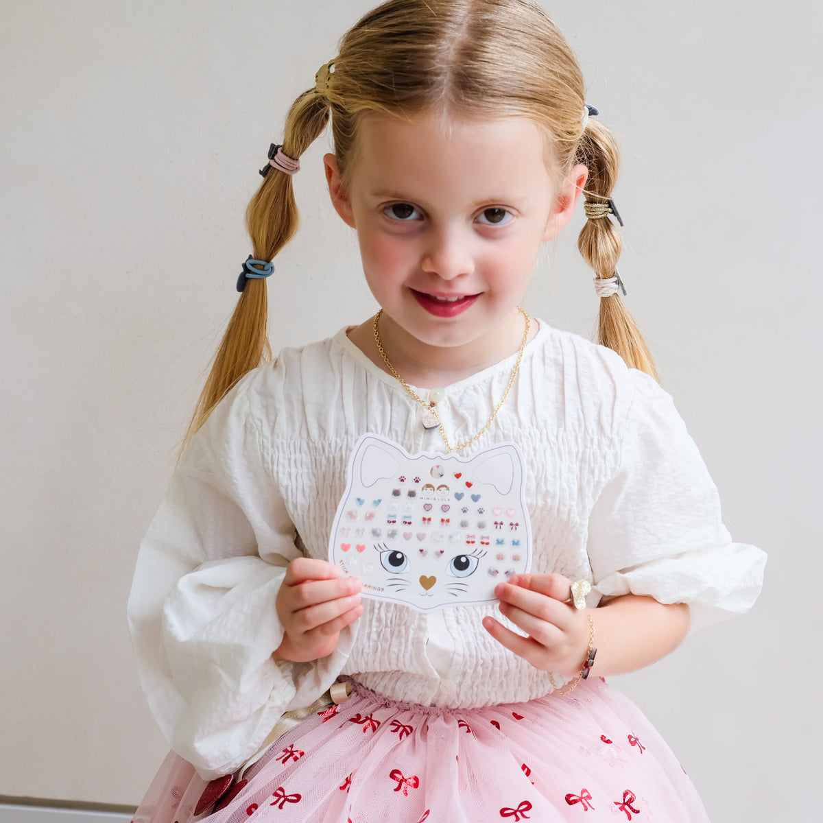 A young girl with blonde pigtails holds a white cat-shaped sticker sheet decorated with colorful hearts, stars, and a gold heart. She is wearing a white long-sleeve blouse and a pink tutu skirt with red bow patterns, set against a plain light gray background.