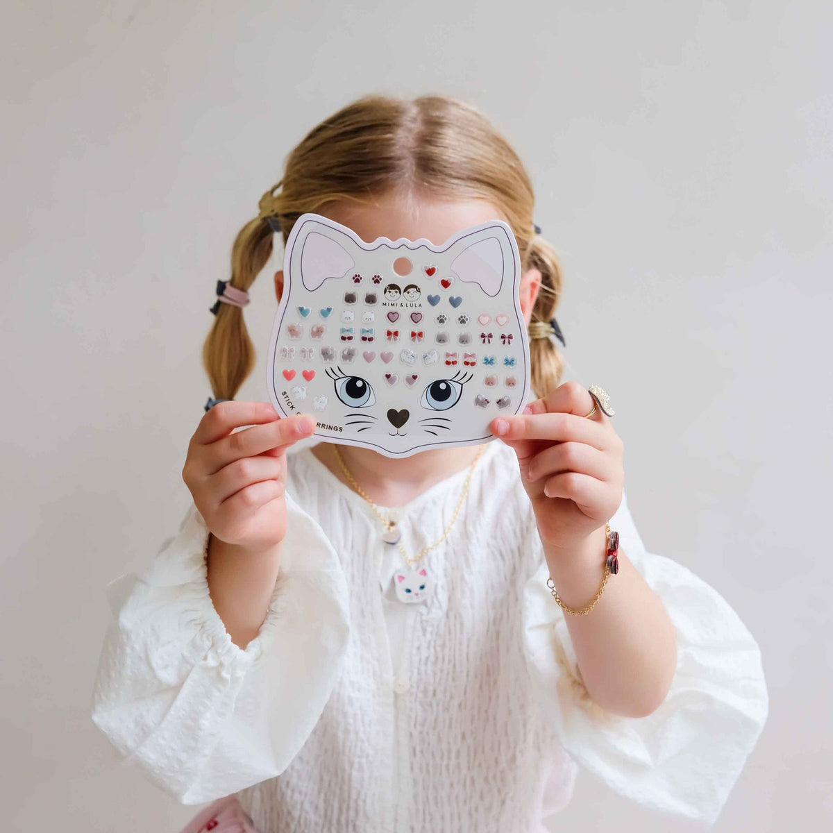 A young child with blonde pigtails holds a white cat-shaped sticker sheet in front of their face, wearing a white blouse against a plain light background.