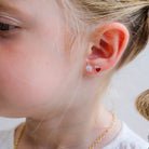 A close-up of a child's ear wearing two small stud earrings: one white with a black face detail and one red heart-shaped stud.