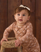 A baby girl wearing a long-sleeve tan jumpsuit with a white floral pattern and a matching cream bow headband, leaning on a rustic wooden stool against a dark wood plank background.