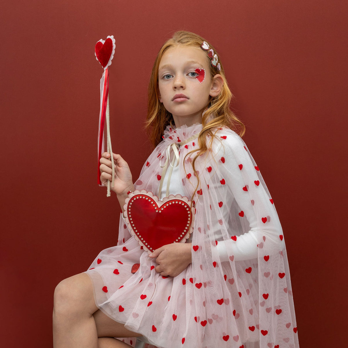 Young girl in a heart-patterned dress holding a red heart-shaped object against a red background
