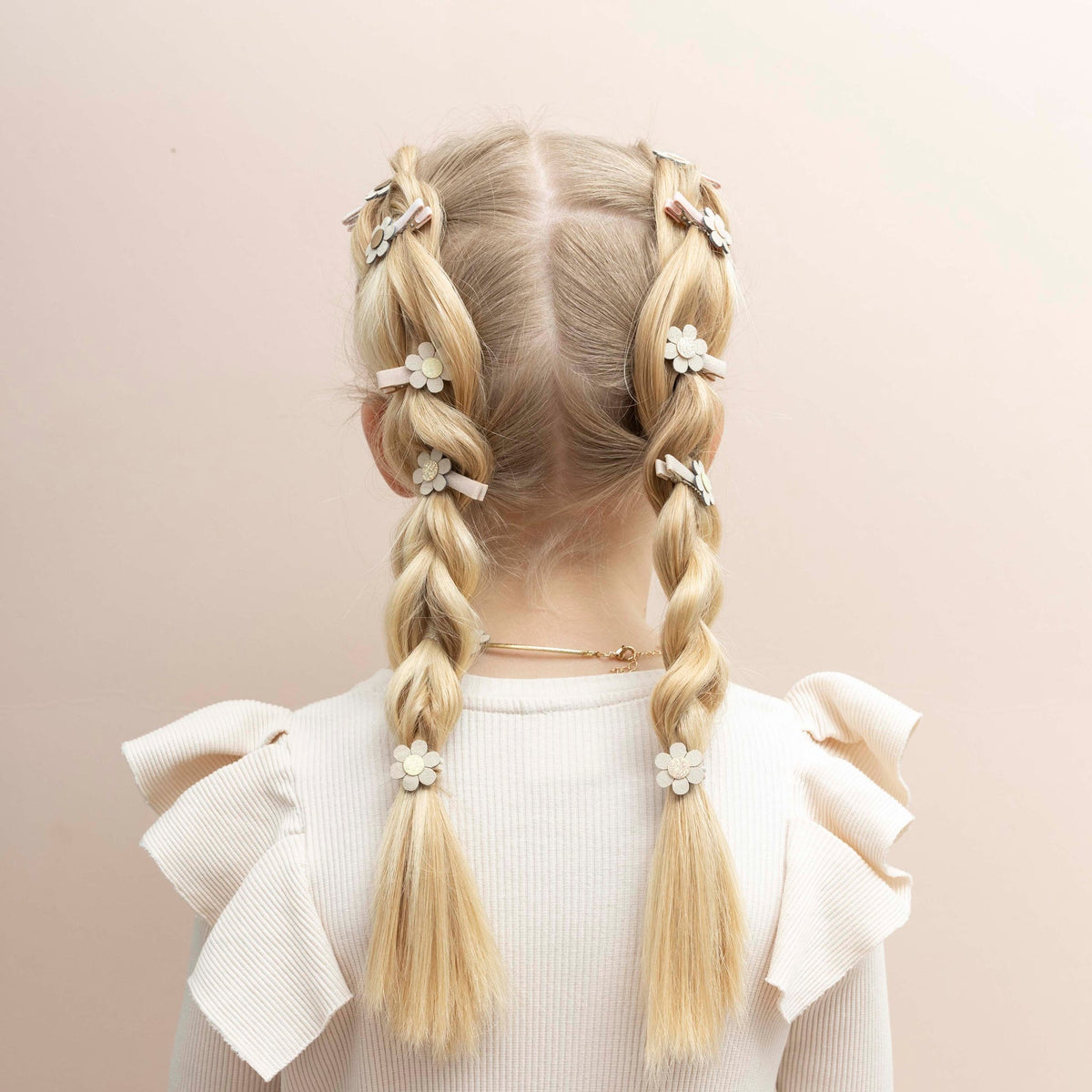 Back view of a child with blonde hair styled in braids, wearing a white top with ruffled sleeves on a beige background.