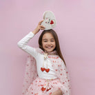 Young girl wearing a white shirt with red hearts and a pink dress, holding a white sandal with cartoon character on a pink background.