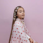 A young girl with long brown hair decorated with heart-shaped hair clips, wearing a white tulle dress with a red bow pattern, posed against a solid pink background.