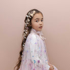 A young girl with long brown hair styled in a braid decorated with white hair bows, wearing a white long-sleeve tulle dress with pastel star and moon prints, posed against a solid pale pink background.