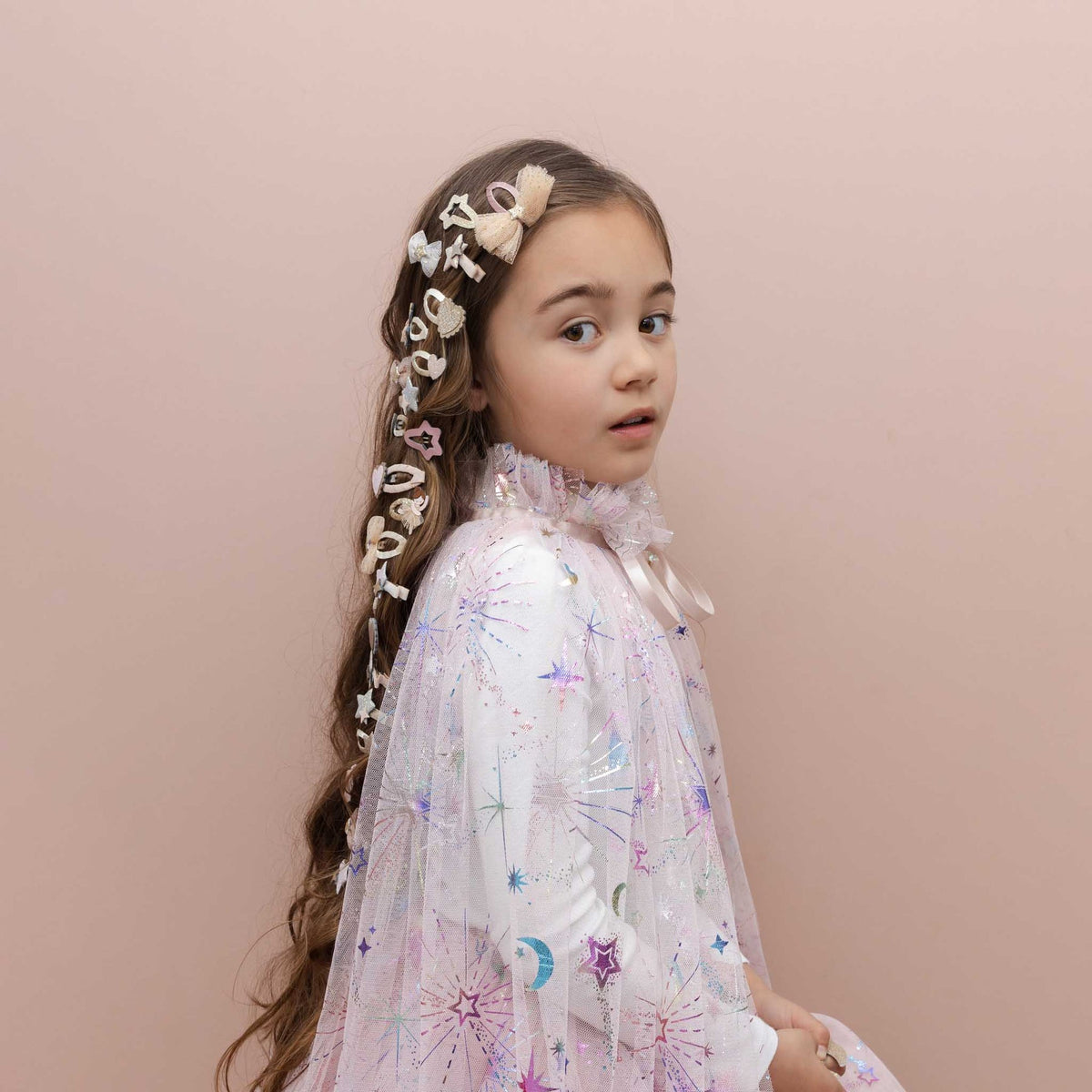 A young girl with long brown hair styled in a braid decorated with white hair bows, wearing a white long-sleeve tulle dress with pastel star and moon prints, posed against a solid pale pink background.