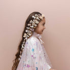 A young girl with long brown hair is shown in profile wearing a decorative headpiece made of multiple white and beige hair clips and bows, along with a sparkly white dress, against a solid pale pink background.