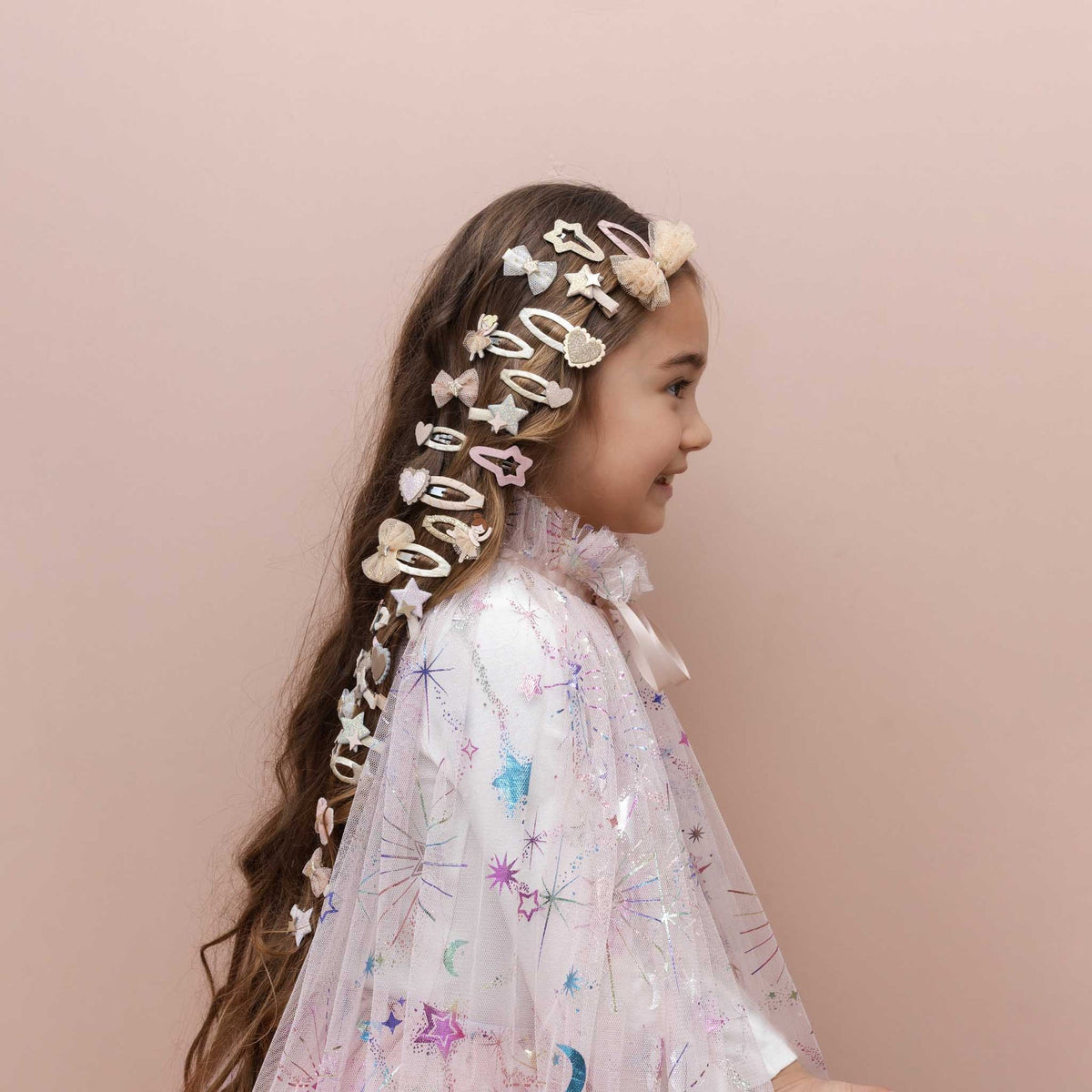 A young girl with long brown hair is shown in profile wearing a decorative headpiece made of multiple white and beige hair clips and bows, along with a sparkly white dress, against a solid pale pink background.