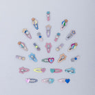 A set of small decorative hair clips arranged in a loose circle on a light gray background. The clips feature pastel embroidered shapes including stars, hearts, and wand-like motifs in pink, blue, purple, yellow, and peach, with silver metal spring clips.