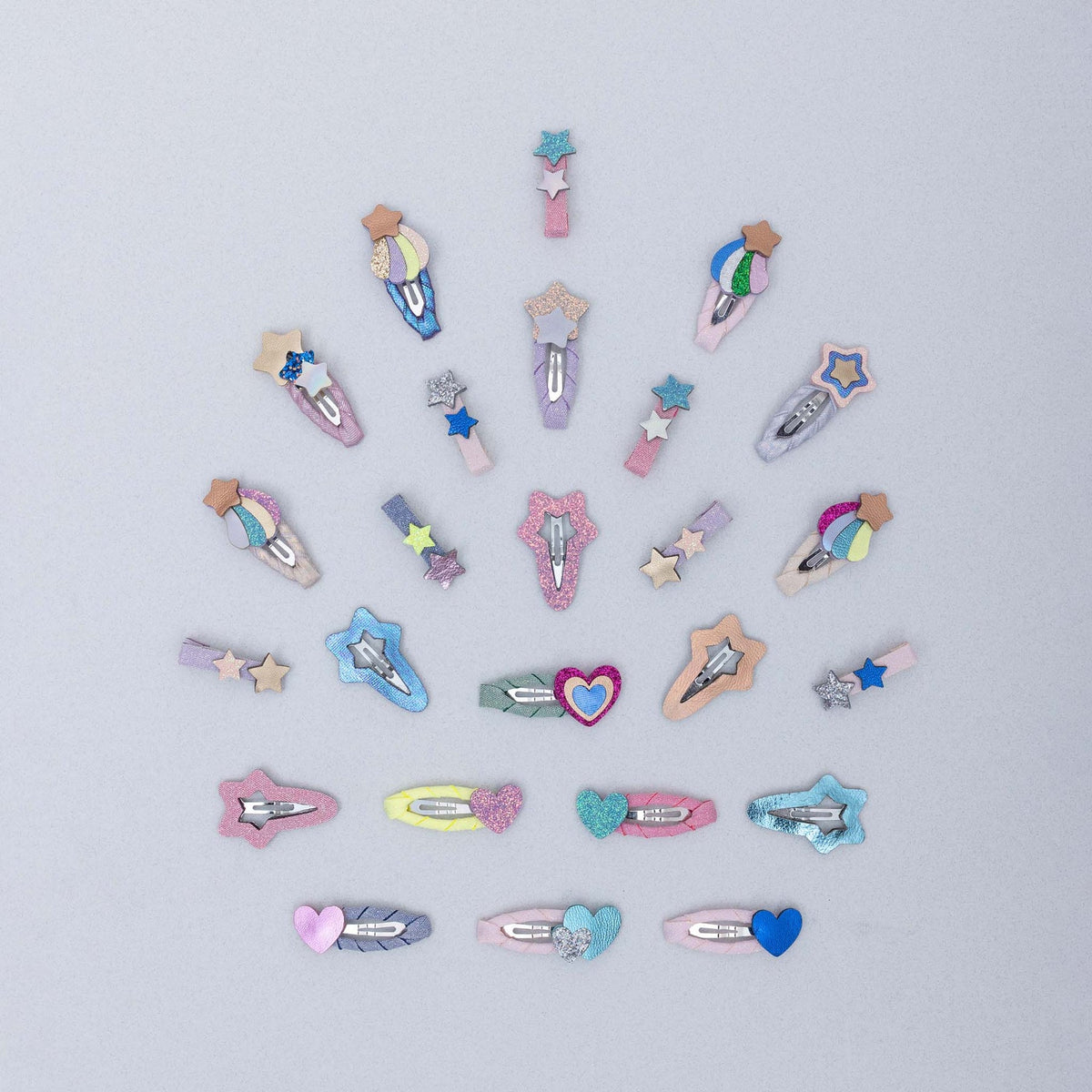 A set of small decorative hair clips arranged in a loose circle on a light gray background. The clips feature pastel embroidered shapes including stars, hearts, and wand-like motifs in pink, blue, purple, yellow, and peach, with silver metal spring clips.