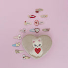 Heart-shaped pillow with cat design surrounded by various small hair accessories on a pink background