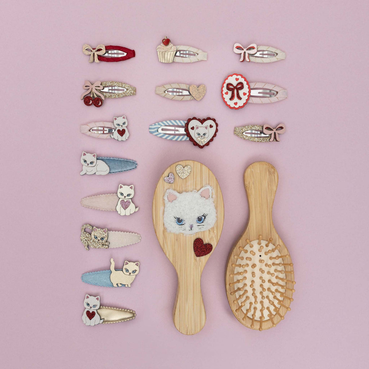 A set of wooden hairbrushes and matching hair clips decorated with cute cat and heart motifs, arranged on a solid pink background.