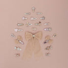 A collection of pastel pink and beige hair clips, bows, and barrettes arranged in a tree-like structure with a large glittery bow at the center, set against a solid dusty pink background.