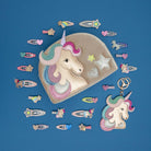 A unicorn-themed hair accessory set arranged on a blue background, including a beige fabric pouch with a unicorn face, matching hair clips, and a keychain.