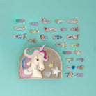 A pastel unicorn-themed hair accessory set arranged on a light aqua background, featuring a large fabric pouch with an embroidered unicorn head and a row of matching hair clips and pins in unicorn, star, and rainbow designs.