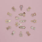A set of 16 pastel hair clips arranged in a circle on a solid pink background. The clips feature whimsical designs including fairies, ballerinas, stars, bows, and hearts in soft pink, beige, and white tones with glittery and metallic details.