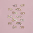 A set of 12 pastel hair clips arranged on a solid pink background. The clips feature whimsical designs including ballerina figures, stars, hearts, bows, and glittery accents in soft pink, beige, and white tones.