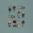 A set of nine handmade hair clips shaped like nature-themed figures, including flowers, a butterfly, a snail, a ladybug, a caterpillar, and a bee, arranged on a solid muted teal background.