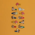 colorful wooden hair clips shaped like nature elements including mushrooms, a squirrel, a hedgehog, a leaf, and a deer, arranged in a grid on a solid orange background.