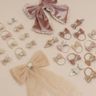A flat-lay collection of pastel hair accessories on a solid beige background, including velvet and tulle bows, hair clips, and hair ties in soft pink, beige, and light blue tones, accented with glittery star details.