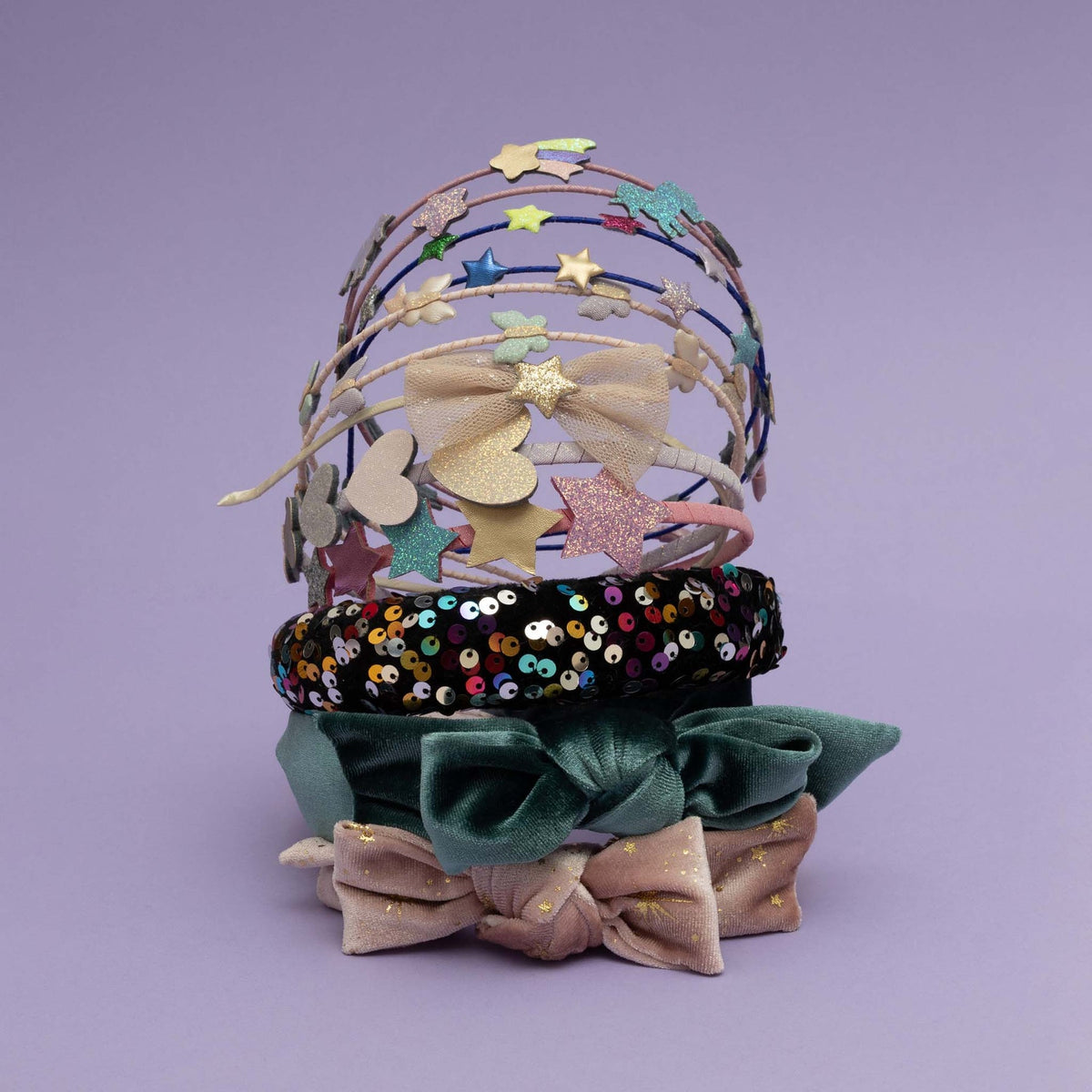 A stack of five decorative headbands on a solid lavender background. The stack includes a gold wire tiara with glittery star and heart accents, a black headband with multicolor sequins, a dark green velvet headband with a bow, a dusty pink headband with gold glitter stars, and a matching pink bow.