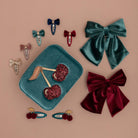 A flat-lay display of hair accessories on a dusty pink background, featuring two large velvet bows (one teal, one burgundy), several smaller bow clips, and cherry-shaped hair clips on a matching teal velvet tray.