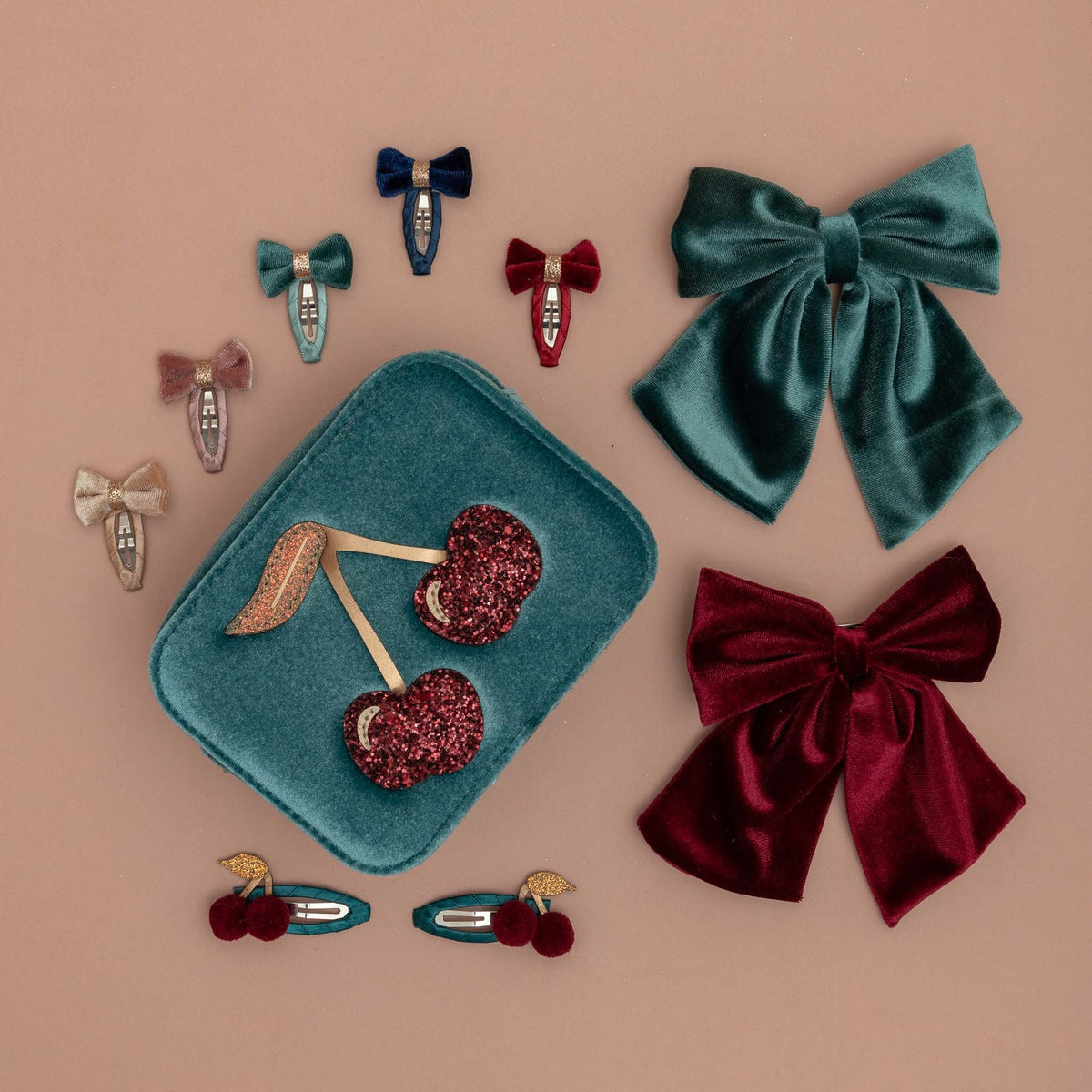 A flat-lay display of hair accessories on a dusty pink background, featuring two large velvet bows (one teal, one burgundy), several smaller bow clips, and cherry-shaped hair clips on a matching teal velvet tray.
