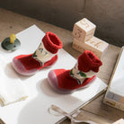 Red baby shoes with white soles on a wooden surface with blocks and a pacifier.