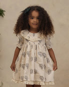A young girl with curly brown hair wearing a cream-colored short-sleeve dress with a blue floral block print and lace trim, standing against a textured beige wall.