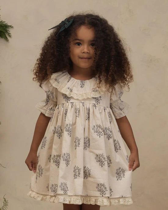 A young girl with curly brown hair wearing a cream-colored short-sleeve dress with a blue floral block print and lace trim, standing against a textured beige wall.