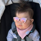 A baby wearing pink mirrored sunglasses with a pink bib and light gray jacket, sitting in a stroller.