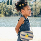 Child with a striped bag featuring a lemon design, standing by a lake.