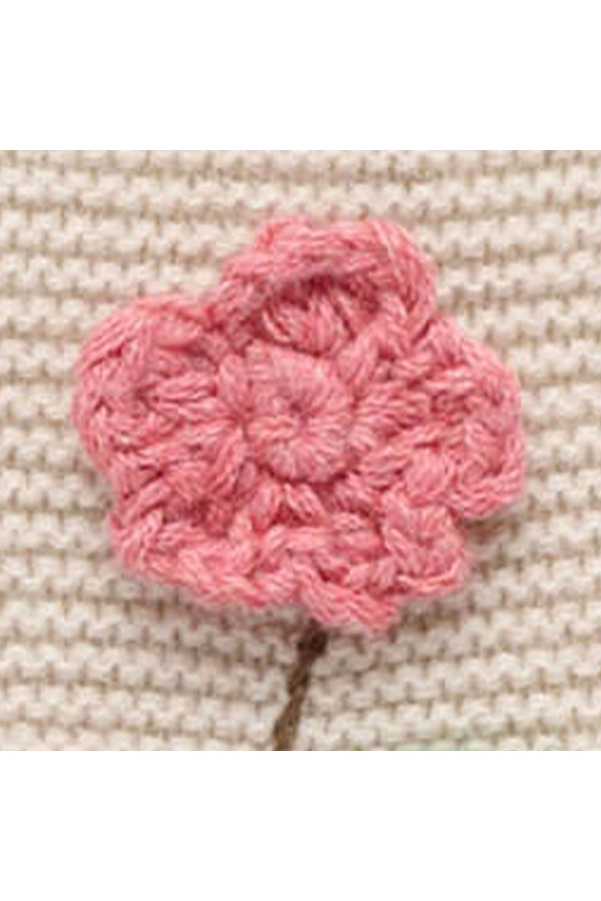 A pink crocheted flower applique with a small brown stem, shown on a textured beige knit fabric background.
