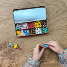 Children's hands working with colorful beads on a wooden table, with a tin of beads open in the background.