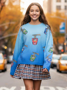 A young girl smiling on a city street, wearing a bright blue crewneck sweater with colorful cartoon noodle cup prints and a brown, white, and blue plaid mini skirt.