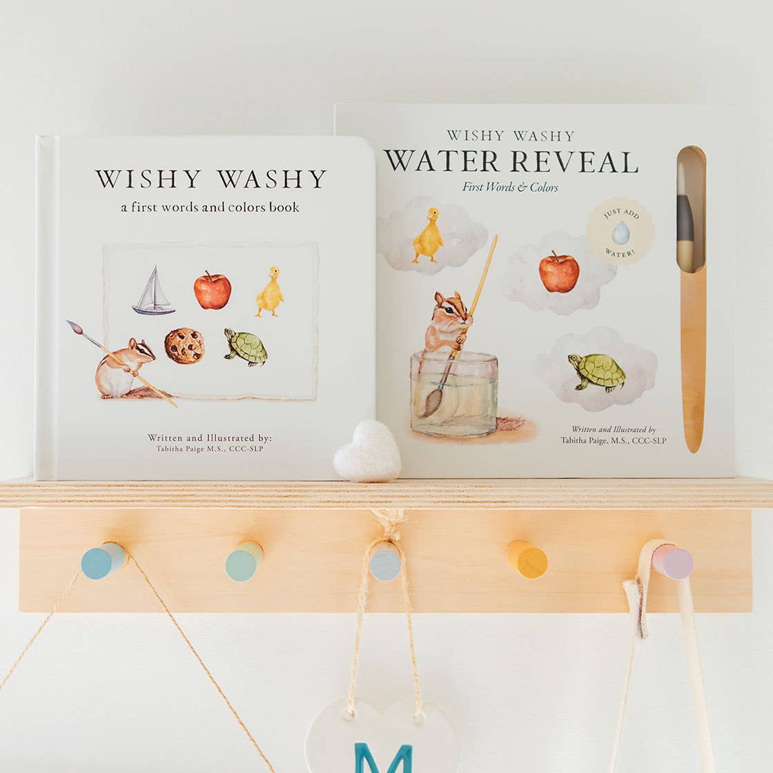 Two children's books displayed on a light wood shelf: 'Wishy Washy' and 'Wishy Washy Water Reveal' with a wooden applicator tool, against a white wall with colorful pegs.