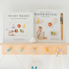 Two children's books displayed on a light wood shelf: 'Wishy Washy' and 'Wishy Washy Water Reveal' with a wooden applicator tool, against a white wall with colorful pegs.