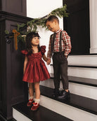 A young girl in a red ruffled dress and red-and-white plaid shirt stands beside a boy in brown suspenders and a matching plaid shirt on a black-and-white staircase, holding hands under a green garland.