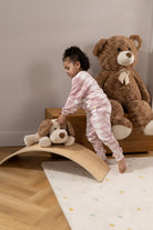 A toddler in a pink and white striped pajama set stands on a wooden arch play ramp in a child's bedroom, holding a plush dog toy. A large brown teddy bear sits on a wooden bench in the background.