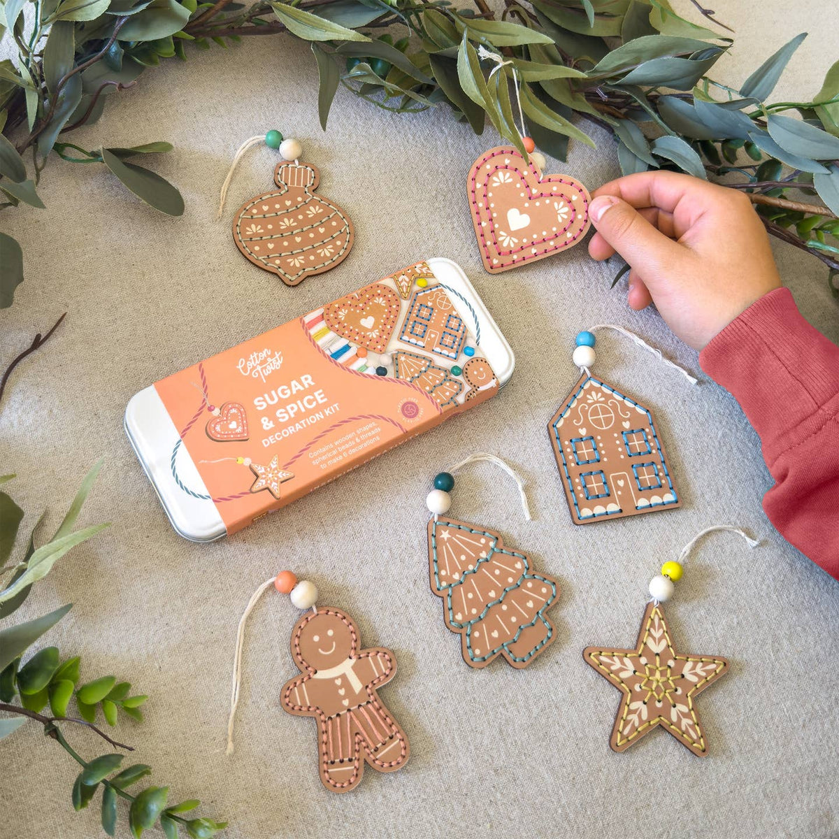 A hand holds a gingerbread heart ornament next to a tin of Sugar & Spice Sugar Cookies, surrounded by five decorated gingerbread cookies shaped like a house, tree, gingerbread man, star, and heart, styled with green eucalyptus leaves on a neutral background.