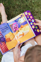 A child holding open "What Does Ñ Sound Like?: A Spanish-English Sound Book," showing an interior illustrated page featuring a purple animated character, a living room scene, and the text "la ARAÑA" on the left, with a vertical panel of sound buttons on the right edge.
