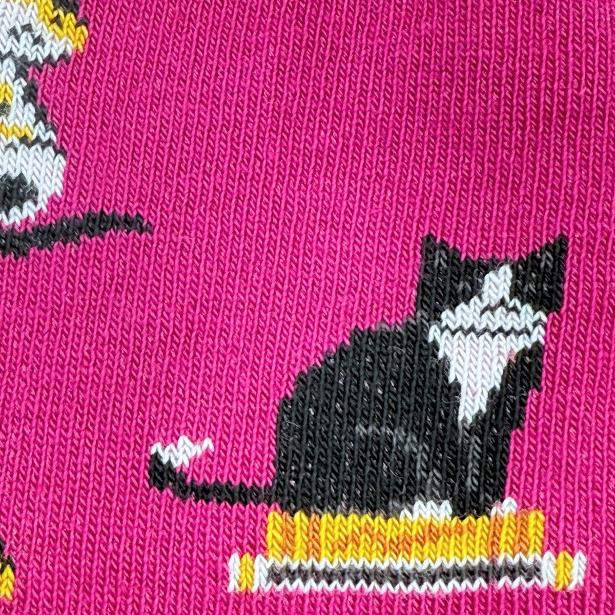 A close-up of a bright pink knit fabric featuring a black-and-white cat sitting on a yellow and white striped base.