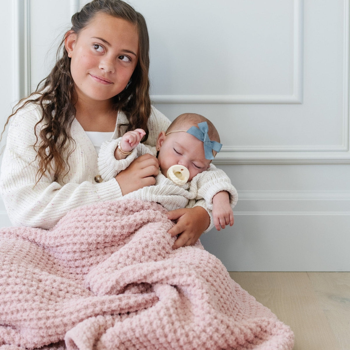 A woman holding a sleeping newborn baby wrapped in a chunky pink blanket. The woman wears a cream knit cardigan, and the baby wears a matching cream outfit with a light blue bow headband.