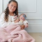 A woman holding a sleeping newborn baby wrapped in a chunky pink blanket. The woman wears a cream knit cardigan, and the baby wears a matching cream outfit with a light blue bow headband.