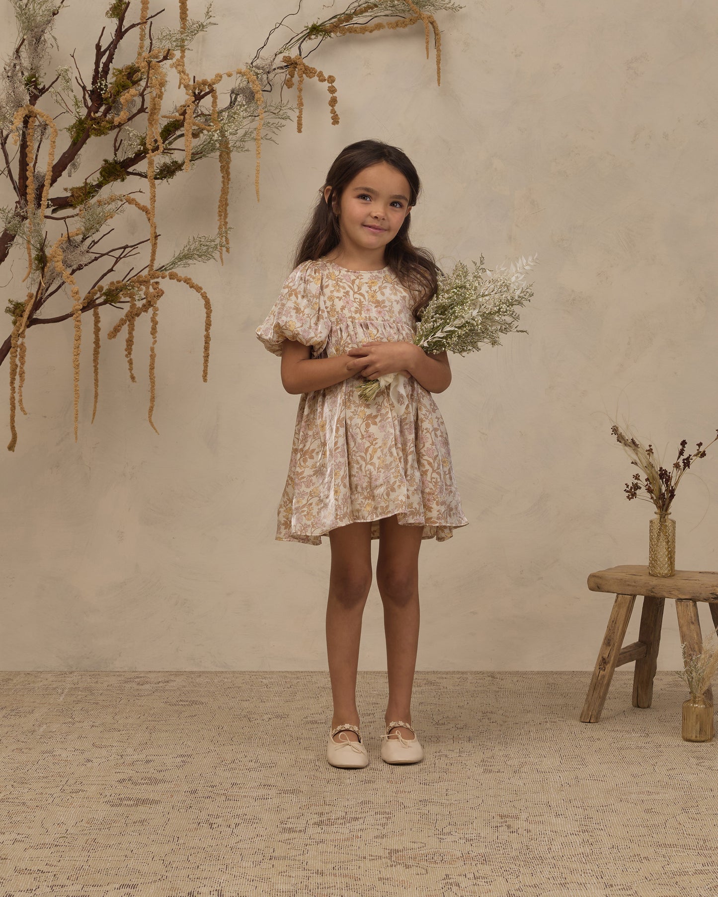 A young girl stands in a studio setting wearing a short, flutter-sleeve dress with a beige and yellow floral pattern. She is holding a small bouquet of dried white flowers and standing next to a rustic wooden stool with a vase of dried branches against a textured beige wall.
