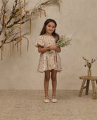 A young girl stands in a studio setting wearing a short, flutter-sleeve dress with a beige and yellow floral pattern. She is holding a small bouquet of dried white flowers and standing next to a rustic wooden stool with a vase of dried branches against a textured beige wall.